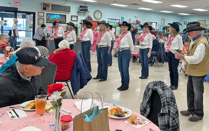 seniors sitting at tables enjoying lunch while performers in black pants, white shirts, and pink scarves perform at CASA Prescott Valley's lunch area